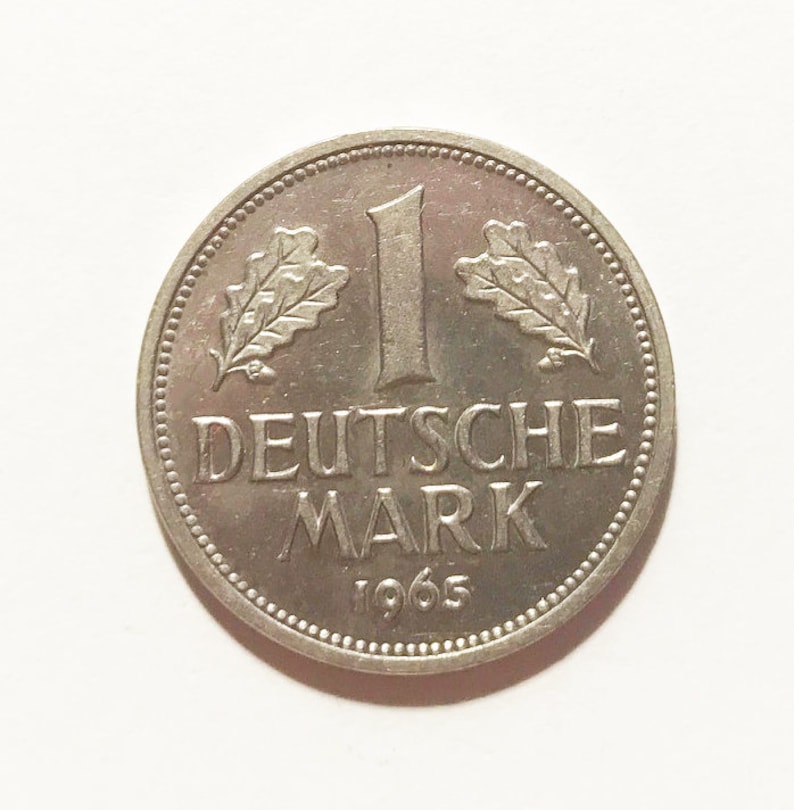 May include: A silver German 1 Deutsche Mark coin from 1965. The coin features an oak leaf design on either side of the number "1" and the words "Deutsche Mark" below.