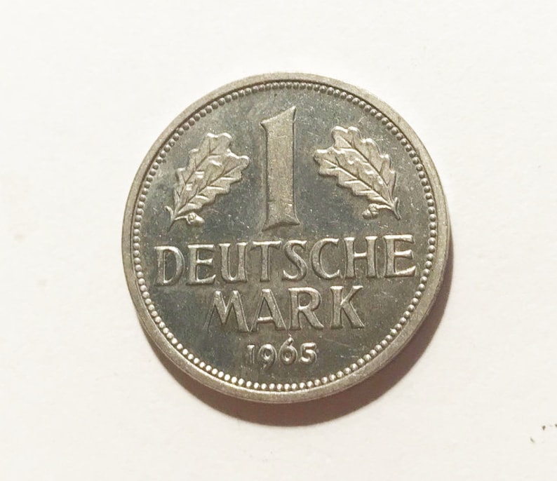 May include: A silver German 1 Deutsche Mark coin from 1965. The coin features an oak leaf on each side of the number "1" and the words "Deutsche Mark" below.