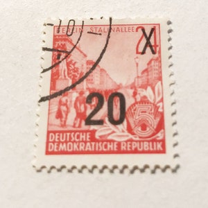 May include: A red and white stamp from the Deutsche Demokratische Republik (German Democratic Republic) with the text "Berlin Stalinallee" and "20" in black. The stamp features a red image of a street scene with buildings and people.
