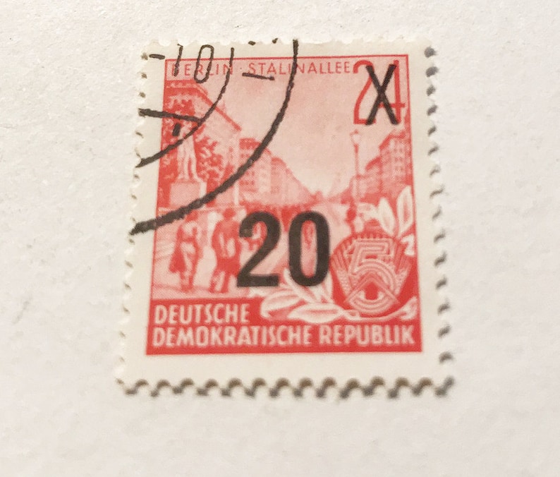 May include: A red and white stamp from the Deutsche Demokratische Republik (German Democratic Republic) with the text "Berlin Stalinallee" and "20" in black. The stamp features a red image of a street scene with buildings and people.