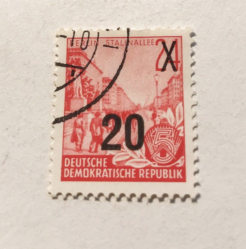 May include: A red and white stamp from the German Democratic Republic with the text "Deutsche Demokratische Republik" and the number "20" printed in black. The stamp features a red image of a street scene with buildings and people walking. The stamp is postmarked with a black stamp that says "Berlin Stalinallee" and the number "24".