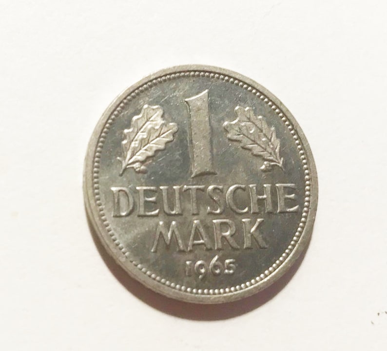 May include: A silver German 1 Deutsche Mark coin from 1965. The coin features an oak leaf on each side of the number "1" and the words "Deutsche Mark".