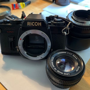Peut inclure: Un appareil photo argentique 35 mm Ricoh KR-10 Super noir avec une bague d'objectif argentée et un objectif Ricoh Rikenon 50 mm f/1.7. L'appareil photo est doté d'une sangle en cuir noir.