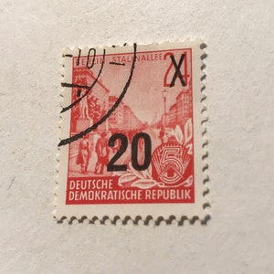 May include: A red and white stamp from the Deutsche Demokratische Republik (German Democratic Republic) with a value of 20. The stamp features a red image of a street scene with a building and people walking. The stamp is postmarked with a black ink stamp that says "Berlin Stalinallee".
