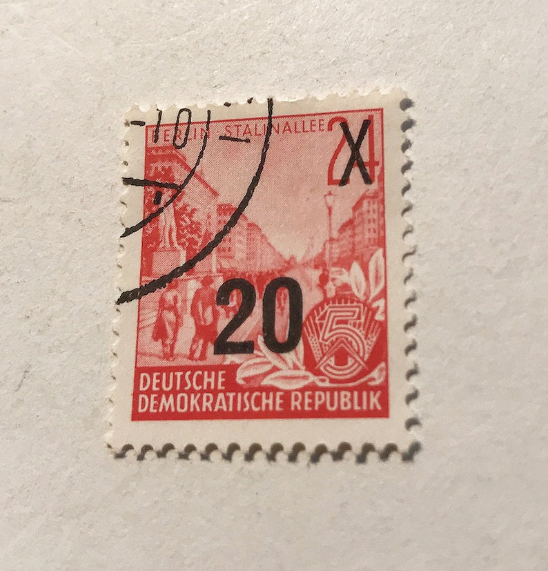 May include: A red and white stamp from the Deutsche Demokratische Republik (German Democratic Republic) with a value of 20. The stamp features a red image of a street scene with a building and people walking. The stamp is postmarked with a black ink stamp that says "Berlin Stalinallee".