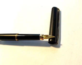 May include: A black fountain pen with a gold trim and a black cap. The pen is lying on a white surface with the cap off.