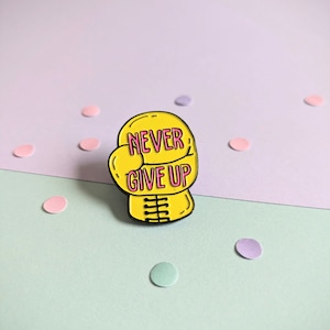 Könnte beinhalten: Gelber Boxhandschuh-Emaille-Pin mit rosa Schriftzug "NEVER GIVE UP".