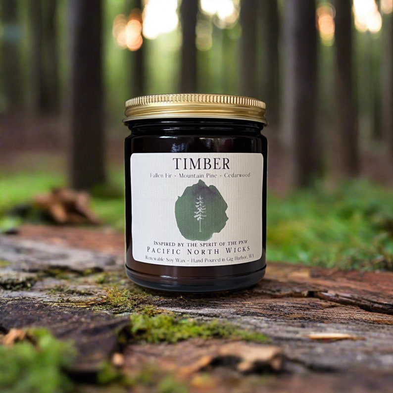 May include: A dark brown glass candle jar with a gold lid. The label reads "TIMBER" and lists the scent as "Fallen Fir - Mountain Pine - Cedarwood". The label also includes the text "INSPIRED BY THE SPIRIT OF THE PNW PACIFIC NORTH WICKS" and "Renewable Soy Wax - Hand Poured in Gig Harbor, WA".