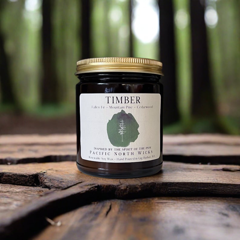May include: A brown glass candle jar with a gold lid. The label reads "TIMBER Fallen Fir - Mountain Pine - Cedarwood" and "INSPIRED BY THE SPIRIT OF THE PNW PACIFIC NORTH WICKS  Renewable Soy Wax - Hand Poured in Gig Harbor, WA".
