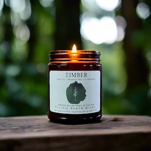 May include: A brown glass candle jar with a white label that reads "TIMBER" and "Fallen Fir - Mountain Pine - Cedarwood". The label also includes the text "INSPIRED BY THE SPIRIT OF THE PNW PACIFIC NORTH WICKS" and "Renewable Soy Wax - Hand Poured in Gig Harbor, WA". The candle is lit and the flame is visible.