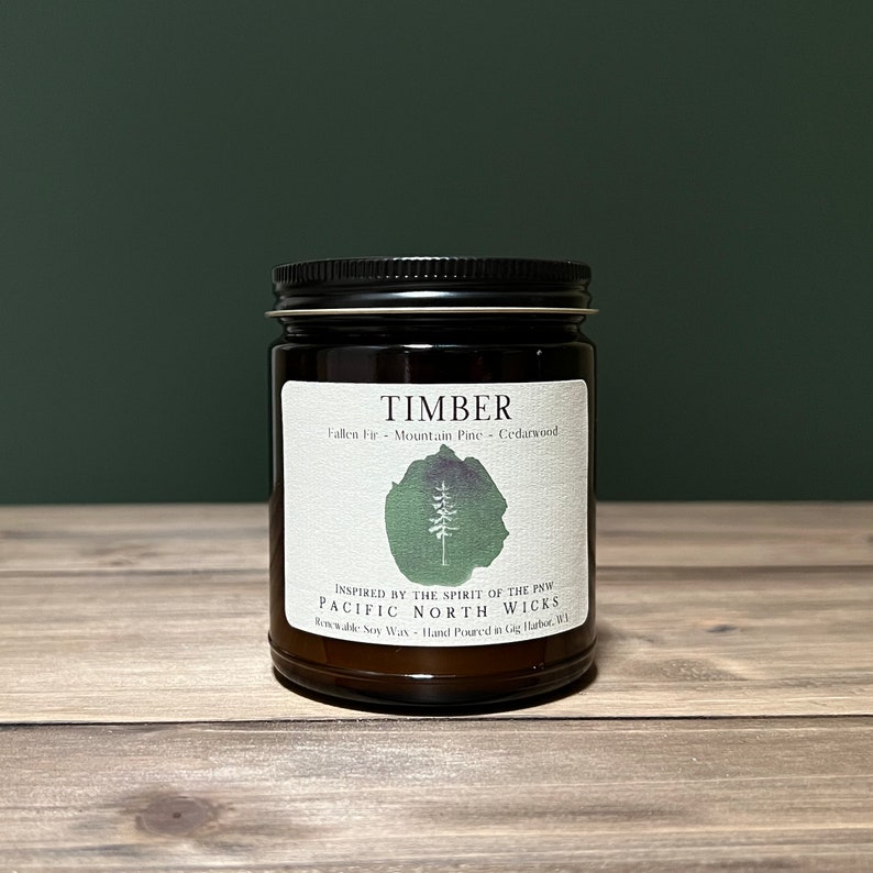May include: A brown glass candle jar with a black lid. The label reads "TIMBER" and lists the scent as "Fallen Fir - Mountain Pine - Cedarwood". The label also includes the text "INSPIRED BY THE SPIRIT OF THE PNW" and "PACIFIC NORTH WICKS".