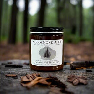 May include: A brown glass candle jar with a black lid and a white label featuring the text WOODSMOKE & FIR, Smoked Embers - Mountain Pine - Cedarwood, and Pacific North Wicks. The label includes a gray watercolor graphic of pine trees.