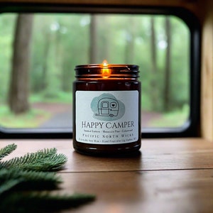 May include: A lit candle in an amber glass jar with a white label featuring a camper illustration and the text HAPPY CAMPER, Smoked Embers - Mountain Pine - Cedarwood, PACIFIC NORTH WICKS, Renewable Soy Wax - Hand Poured in Gig Harbor, WA.