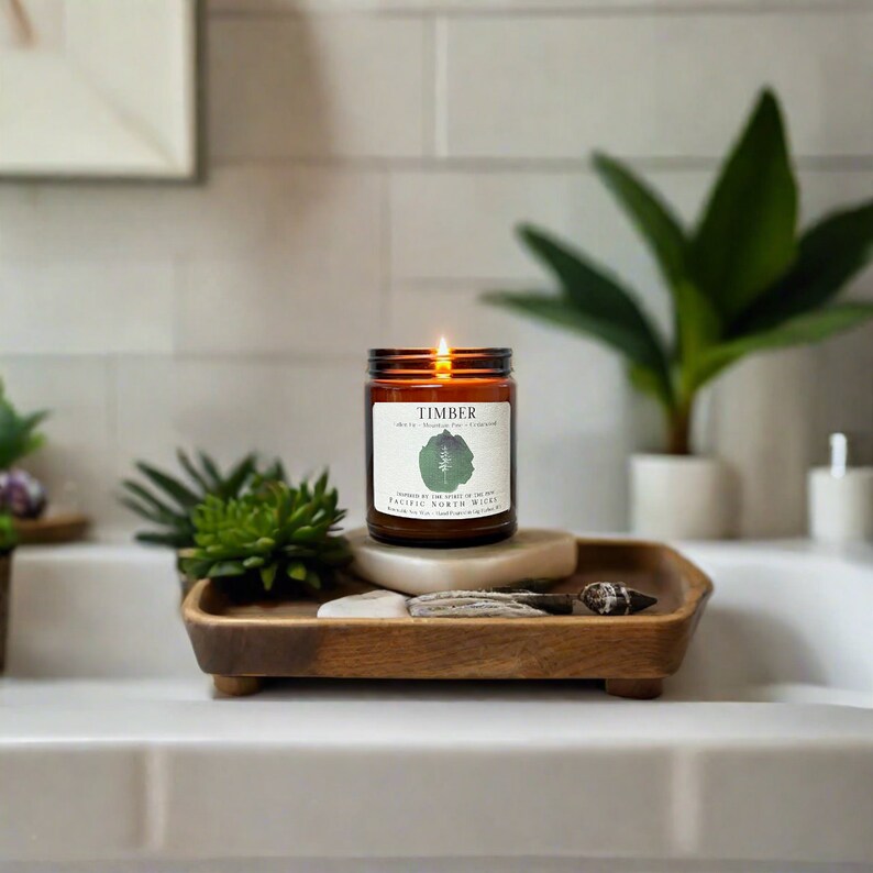May include: A brown glass candle with a white label that reads "TIMBER" and "PACIFIC NORTH WICKS". The candle is lit and sitting on a wooden tray with a white stone and a seashell.
