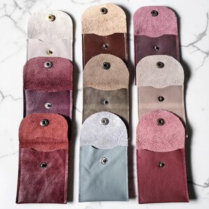 Overhead view of several leather pouches in shades of beige, burgundy, and grey. Each pouch is rectangular with a rounded top flap secured by a snap. The pouches are arranged vertically against a marble background.
