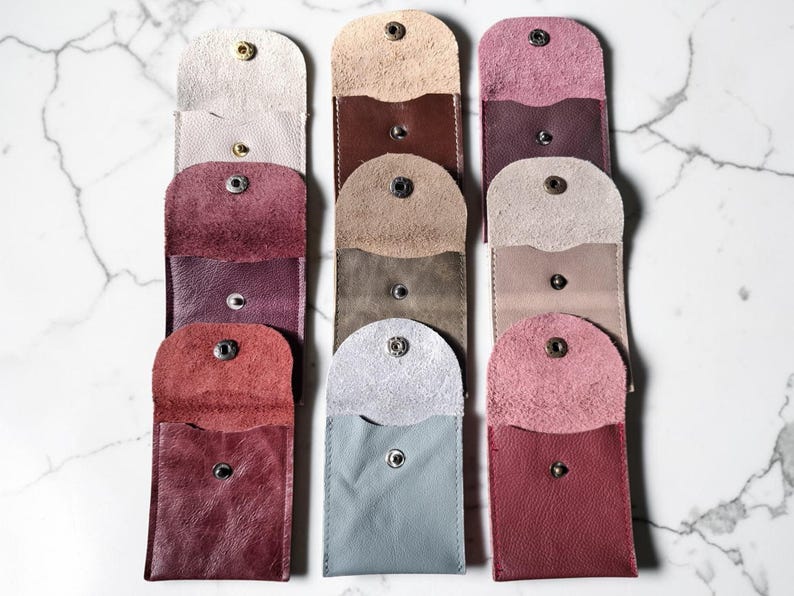 Overhead view of several leather pouches in shades of beige, burgundy, and grey. Each pouch is rectangular with a rounded top flap secured by a snap. The pouches are arranged vertically against a marble background.