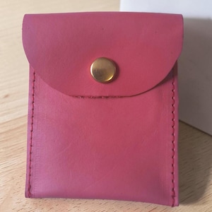 May include: A small, rectangular pink leather pouch featuring a rounded flap closure secured with a single brass-colored metal snap button. The pouch is constructed with visible stitching along the sides and has a smooth, matte finish.