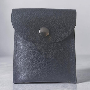 May include: A small, square-shaped dark navy blue pouch with a textured, pebbled surface. The pouch features a flap closure secured by a single, centered, polished silver-toned metal snap button. The edges are stitched with matching dark navy thread.