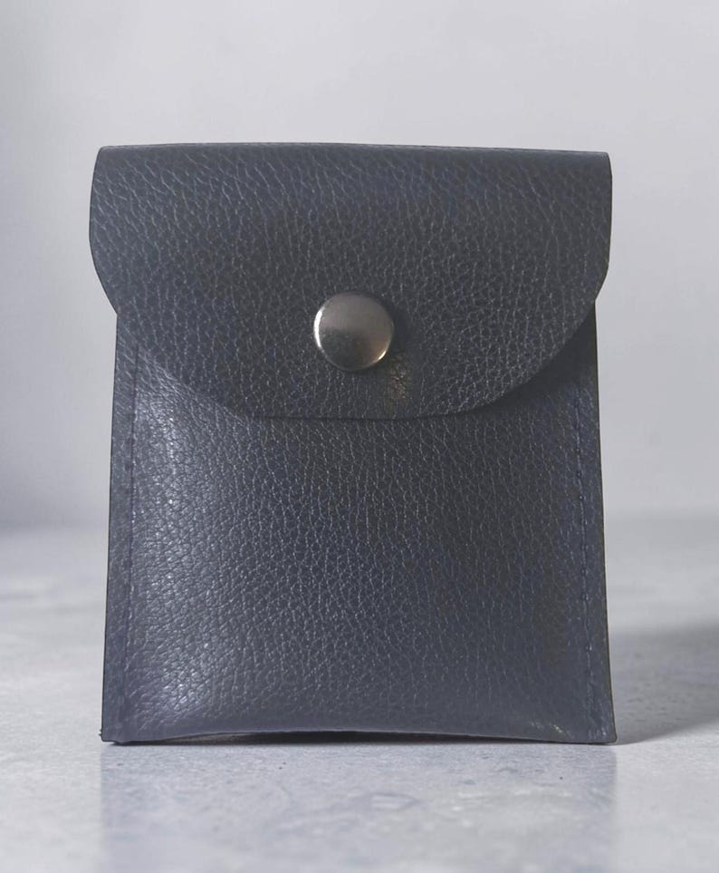 May include: A small, square-shaped dark navy blue pouch with a textured, pebbled surface. The pouch features a flap closure secured by a single, centered, polished silver-toned metal snap button. The edges are stitched with matching dark navy thread.
