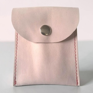 May include: A small, pale pink leather pouch with a rounded flap closure secured by a single silver-toned metal snap button. The sides of the pouch are stitched together with visible red thread, creating a distinct contrast against the light leather.