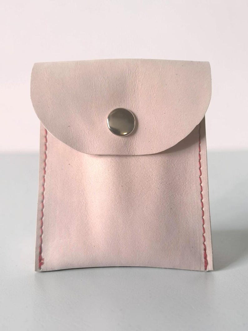 May include: A small, pale pink leather pouch with a rounded flap closure secured by a single silver-toned metal snap button. The sides of the pouch are stitched together with visible red thread, creating a distinct contrast against the light leather.