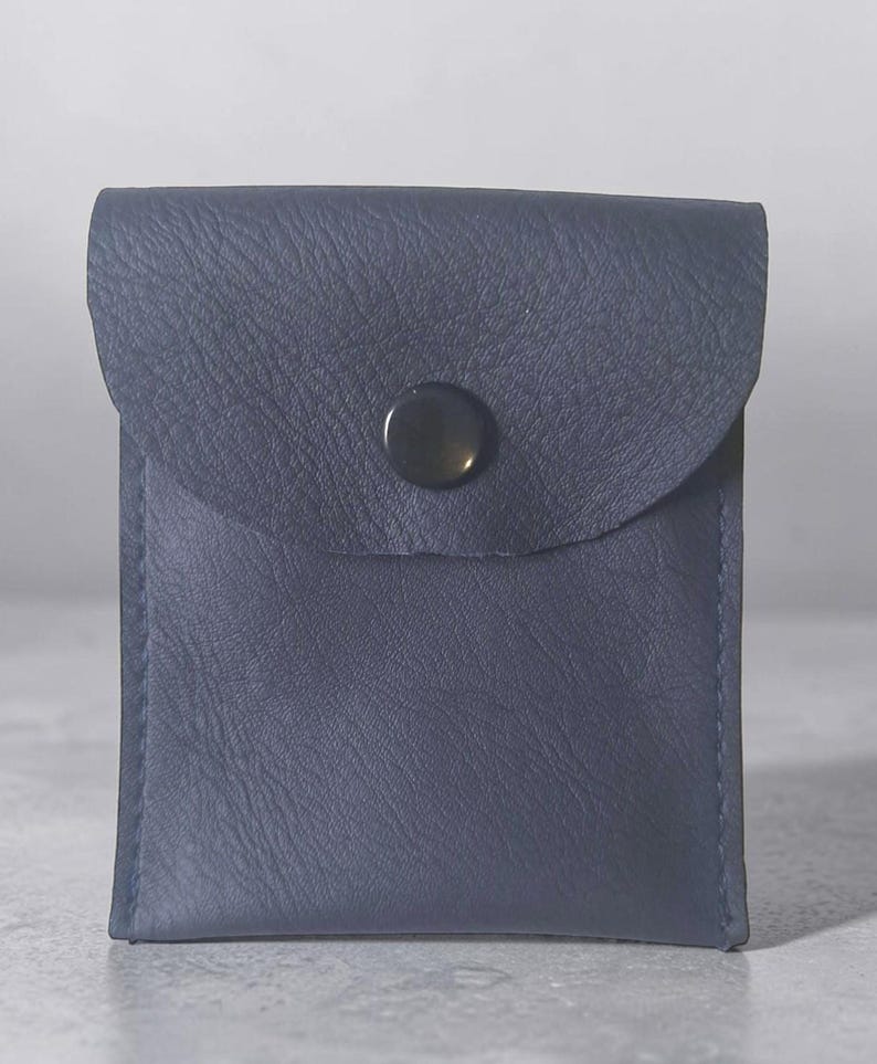 May include: A square-shaped navy blue leather pouch featuring a textured surface and a single centered black snap button closure on the front flap. The pouch has visible stitching along the side edges and a smooth, consistent dark blue color.