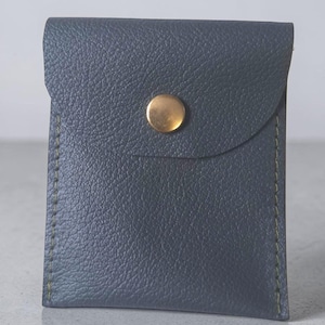May include: A small, rectangular, dark blue pouch with a textured surface and visible green stitching along the sides. The pouch features a flap closure secured by a single, centered, circular brass-colored metal snap button fastener.