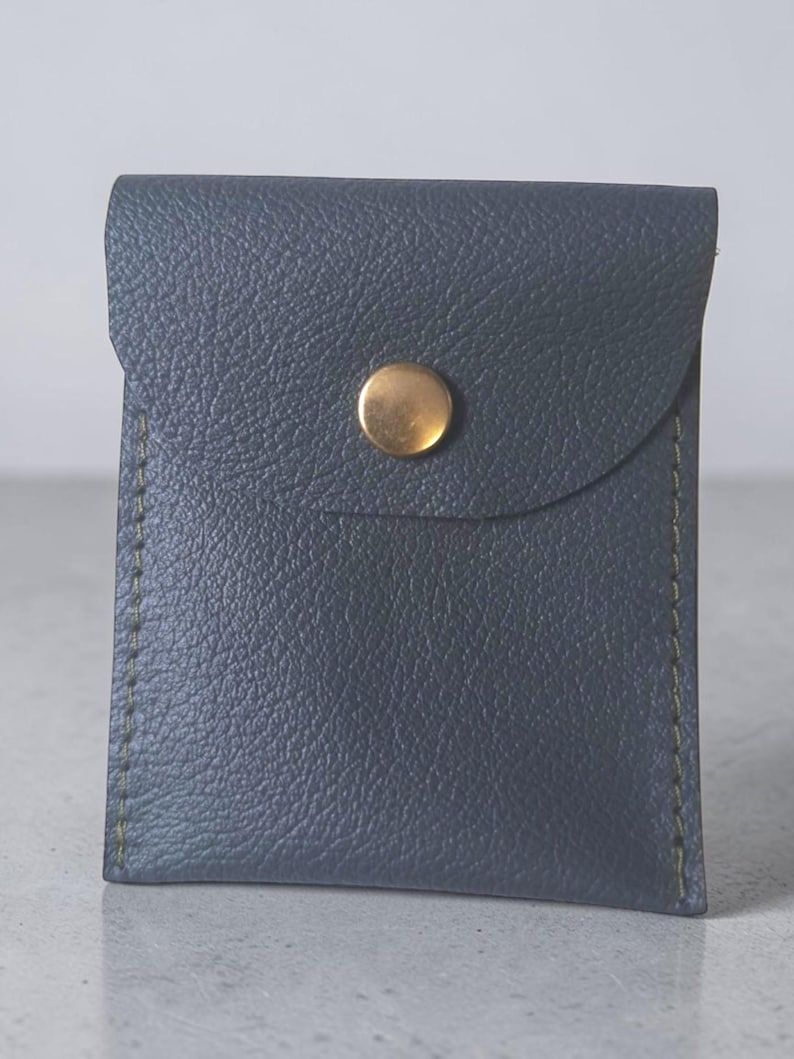 May include: A small, rectangular, dark blue pouch with a textured surface and visible green stitching along the sides. The pouch features a flap closure secured by a single, centered, circular brass-colored metal snap button fastener.