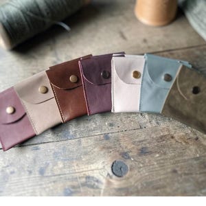 A selection of leather coin purses in shades like burgundy, beige, and olive. Each small wallet has a snap closure. The purses are made of leather.