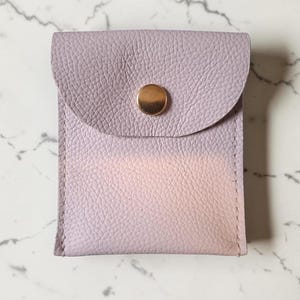 May include: A small, rectangular pouch made of textured, light lavender leather. The pouch features a rounded flap closure secured by a single, centered, polished gold-tone metal snap button. The material has a visible pebbled grain texture.