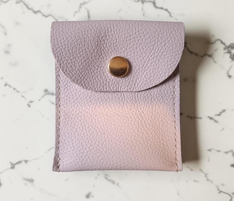 May include: A small, rectangular pouch made of textured, light lavender leather. The pouch features a rounded flap closure secured by a single, centered, polished gold-tone metal snap button. The material has a visible pebbled grain texture.