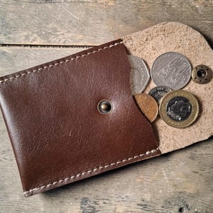A brown leather coin purse, open to show several coins. The purse has a snap closure and is stitched with cream thread. The coins are of different sizes and designs. The purse rests on a wooden surface.