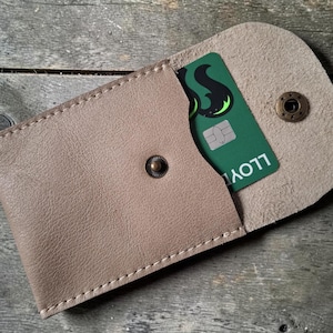 A beige leather cardholder with a press stud closure, containing a green credit card. The cardholder is open, showing the card with the word "LLOYD" printed on it. The cardholder is on a wooden surface.