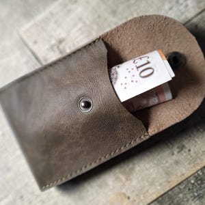 A brown leather coin purse, secured with a snap, with a rolled-up £10 note visible inside. The wallet has a textured finish and is displayed on a wooden background.