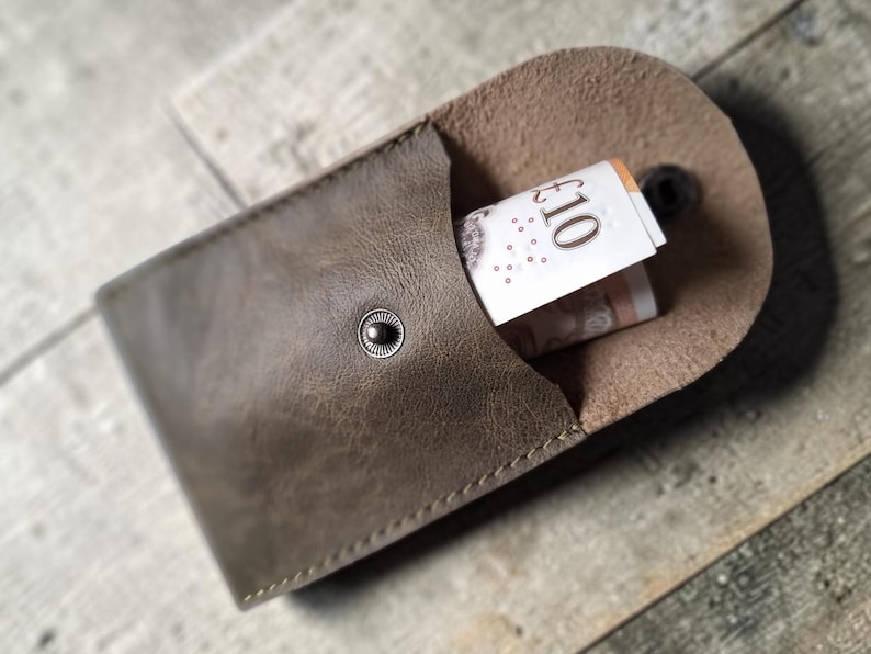 A brown leather coin purse, secured with a snap, with a rolled-up £10 note visible inside. The wallet has a textured finish and is displayed on a wooden background.