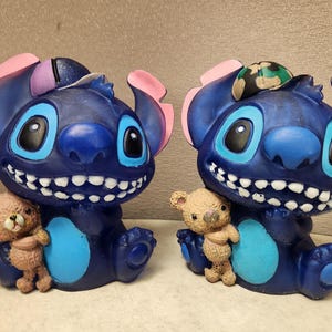 May include: Two blue cartoon character figurines with large smiles, big eyes, and pink ears. Each figurine holds a small brown teddy bear. One has a camouflage accessory on its head. The figurines are sitting down.