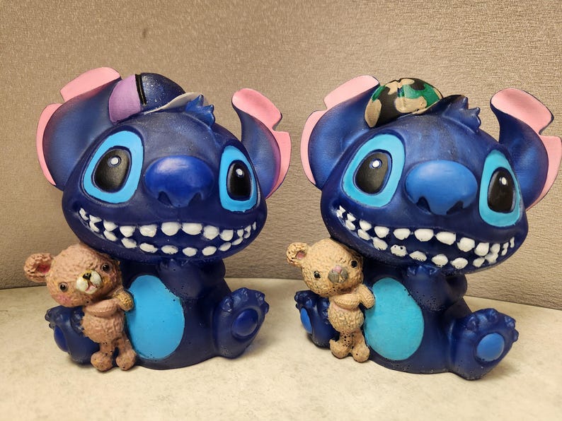 May include: Two blue cartoon character figurines with large smiles, big eyes, and pink ears. Each figurine holds a small brown teddy bear. One has a camouflage accessory on its head. The figurines are sitting down.
