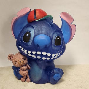 May include: A blue figurine of a cartoon character with large ears, a red and green hat, and a wide grin, holding a small teddy bear. The figurine is painted with details and has a glossy finish.