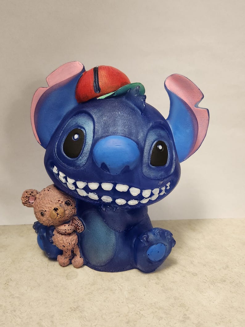 May include: A blue figurine of a cartoon character with large ears, a red and green hat, and a wide grin, holding a small teddy bear. The figurine is painted with details and has a glossy finish.