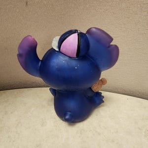 May include: A blue figurine of a cartoon character with large ears and a small teddy bear. The figurine has a purple and pink hat and purple accents on the ears and arms. The figurine is on a white surface.