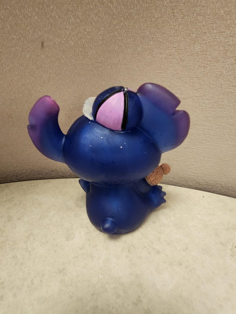 May include: A blue figurine of a cartoon character with large ears and a small teddy bear. The figurine has a purple and pink hat and purple accents on the ears and arms. The figurine is on a white surface.