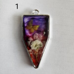 May include: A pendant with a silver-colored frame, featuring a butterfly and a flower encased in resin. The resin has a gradient of colors, including purple, red, and white. The pendant is shaped like a downward-pointing trapezoid.