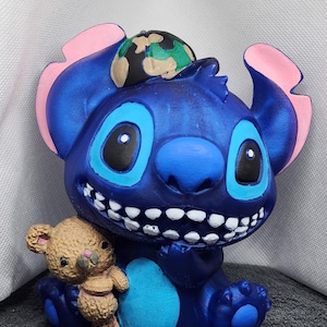 May include: A blue figurine of a cartoon alien character with large eyes, a wide grin, and a small teddy bear. The alien has pink inner ears and a camouflage hat. The teddy bear is light brown. The figurine is a collectible item.
