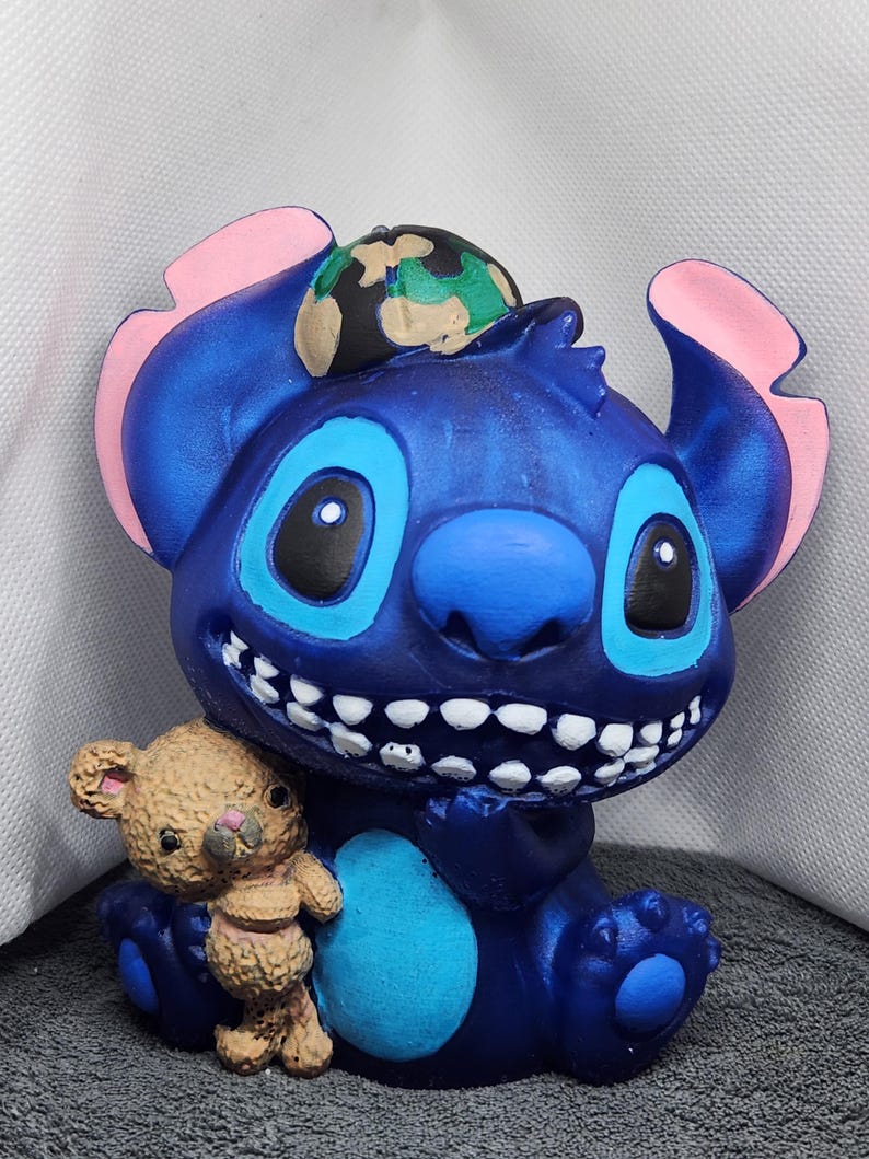May include: A blue figurine of a cartoon alien character with large eyes, a wide grin, and a small teddy bear. The alien has pink inner ears and a camouflage hat. The teddy bear is light brown. The figurine is a collectible item.