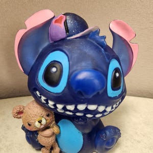 May include: A blue figurine of the character Stitch, with large blue eyes and a wide grin, holding a small teddy bear. The figurine has pink and purple accents on its ears and head. The teddy bear is light brown.