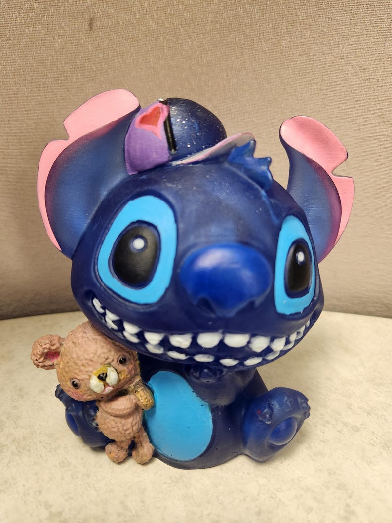 May include: A blue figurine of the character Stitch, with large blue eyes and a wide grin, holding a small teddy bear. The figurine has pink and purple accents on its ears and head. The teddy bear is light brown.