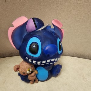 May include: A blue figurine of the character Stitch from Lilo & Stitch, with pink and purple accents on the ears. Stitch is holding a small brown teddy bear. The figurine has large blue eyes and a wide smile.