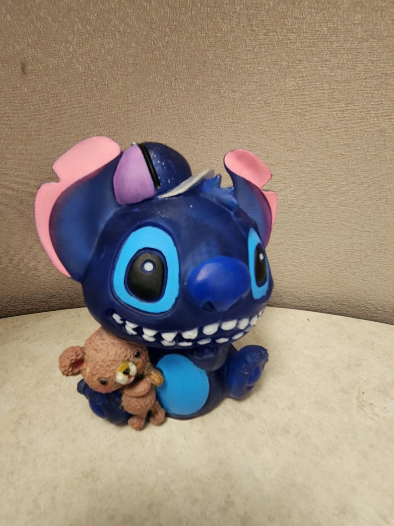 May include: A blue figurine of the character Stitch from Lilo & Stitch, with pink and purple accents on the ears. Stitch is holding a small brown teddy bear. The figurine has large blue eyes and a wide smile.