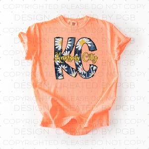 May include: A peach-colored t-shirt with a graphic design of the letters "KC" in a denim fabric with white daisies. The letters are outlined in yellow and the text "Kansas City" is written in yellow below the letters.