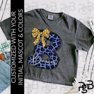 Puede incluir: Camiseta gris oscuro con una gran letra "B" con estampado de leopardo azul y la palabra "Braves" en escritura blanca. Un lazo amarillo con estampado de leopardo está en la parte superior de la letra. La camiseta tiene el texto "PERSONALIZADO CON TU INICIAL, MASCOTA Y COLORES" en el lado izquierdo.
